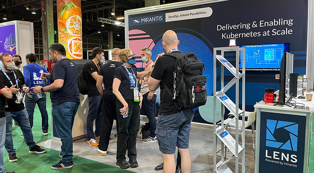 A bustling technology conference booth with MIRANTIS and LENS Powered by Mirantis signage showcases information about Delivering & Enabling Kubernetes at Scale on a large screen as several masked and unmasked individuals interact and engage with the display and booth personnel.